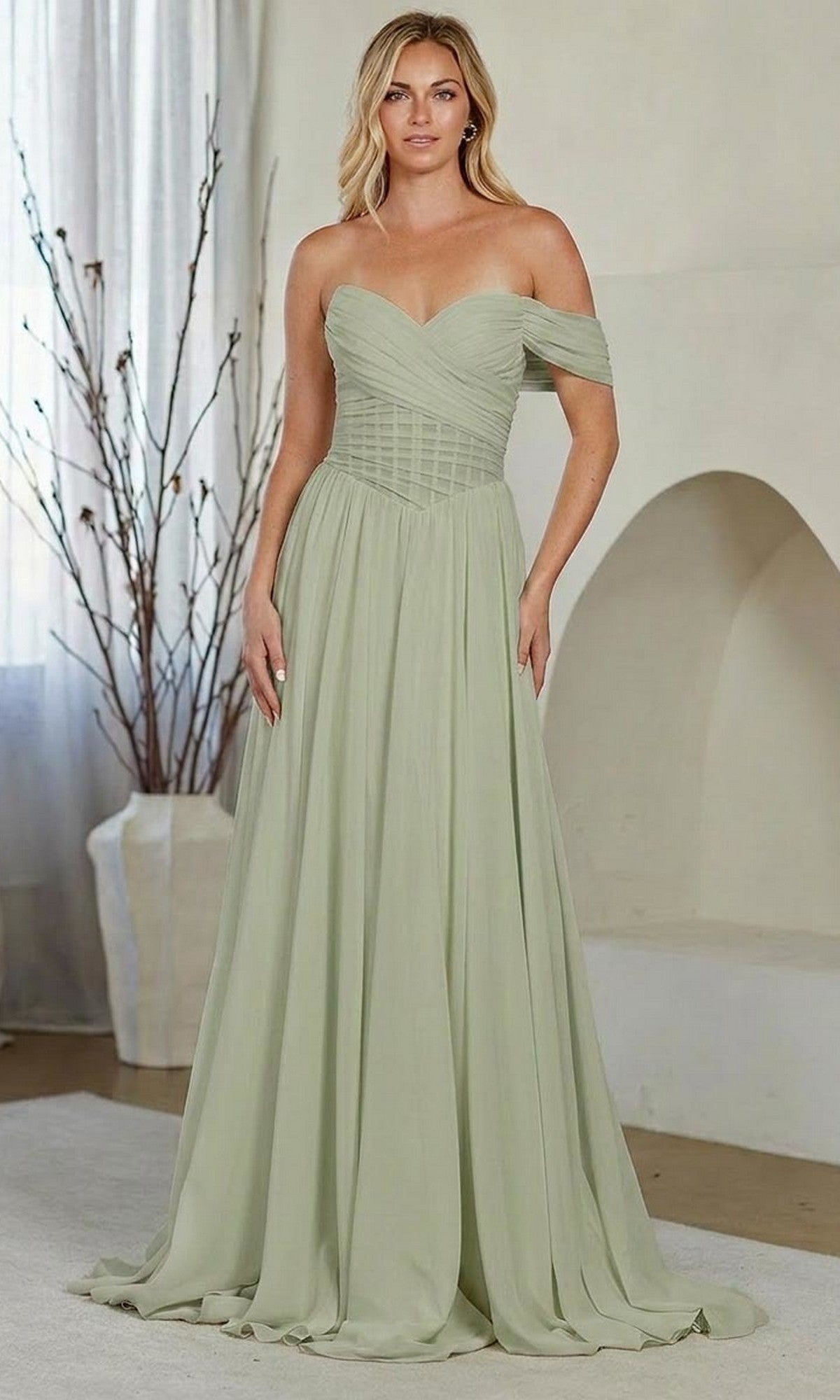 A woman stands indoors in front of a white wall and a vase with branches, wearing the Amelia Couture 9097 long prom dress in sage with an off-the-shoulder neckline and A-line skirt.